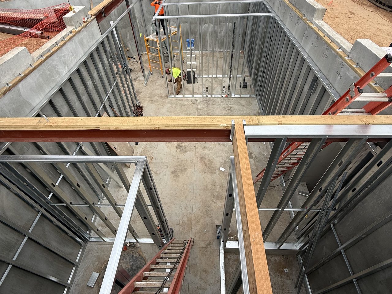 Concrete substructure walls and light gauge steel framing on a below-grade level, with crews working in the pit